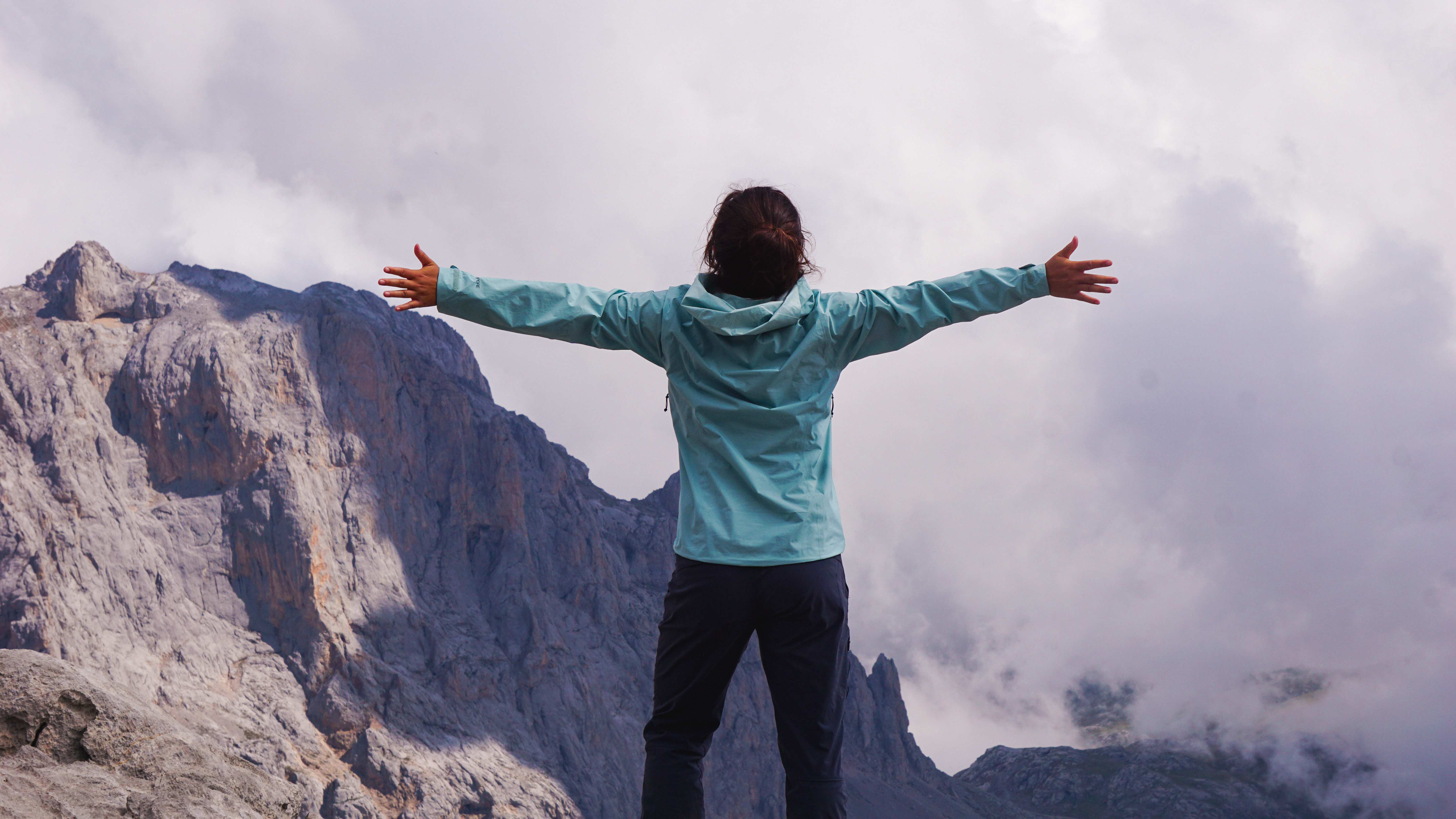 A person standing on a mountain summit with arms outstretched, facing rugged peaks and clouds