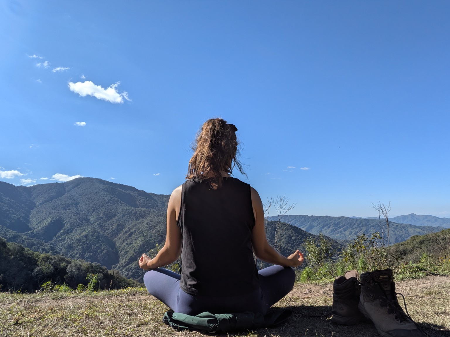 A person sitting cross-legged in meditation on a hilltop, looking out over green forested mountains.