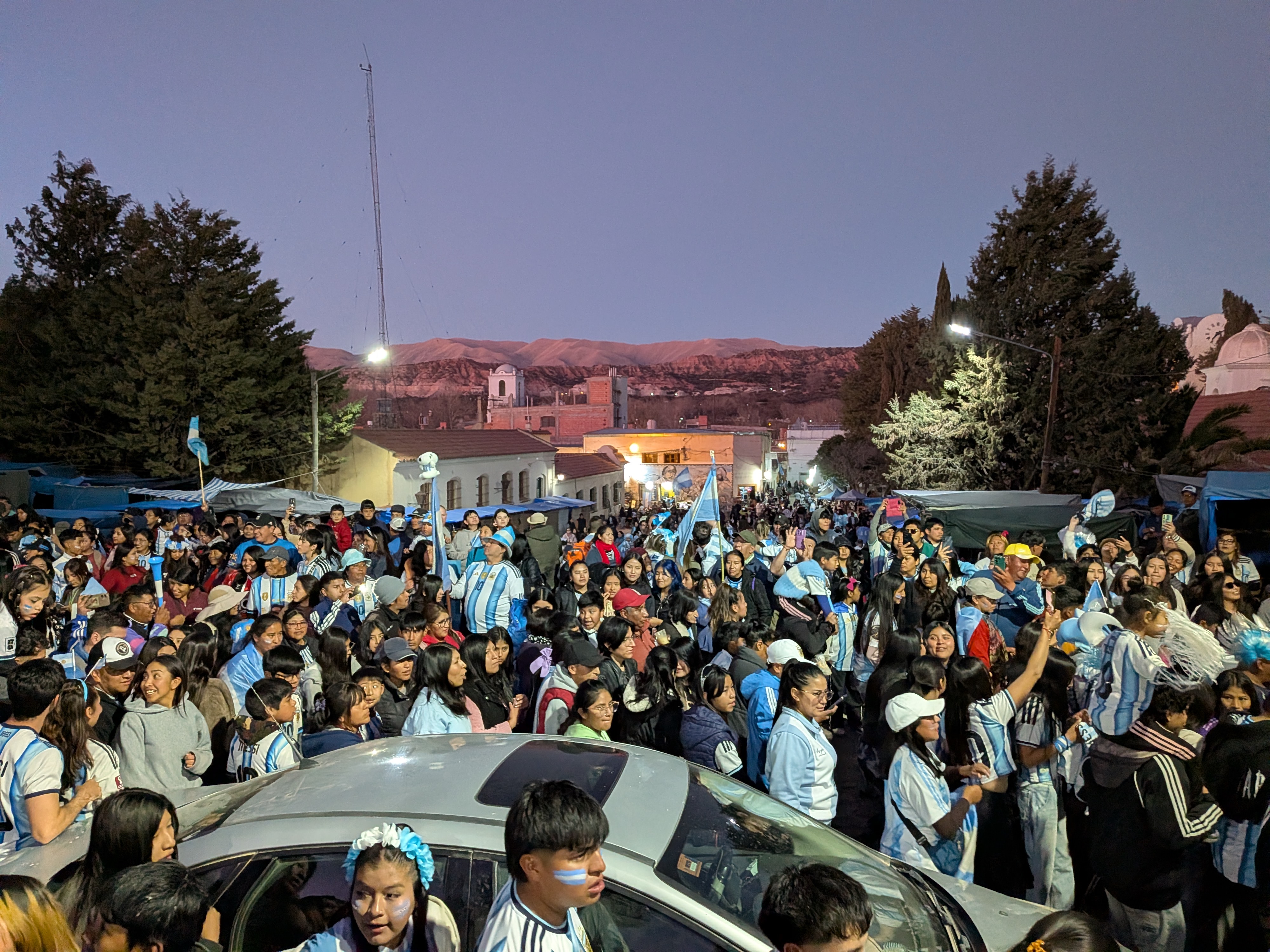 A large evening crowd celebrating in the centre of Humahuaca, dressed in Argentina’s blue-and-white colours and waving flags, with the mountains glowing behind the town.