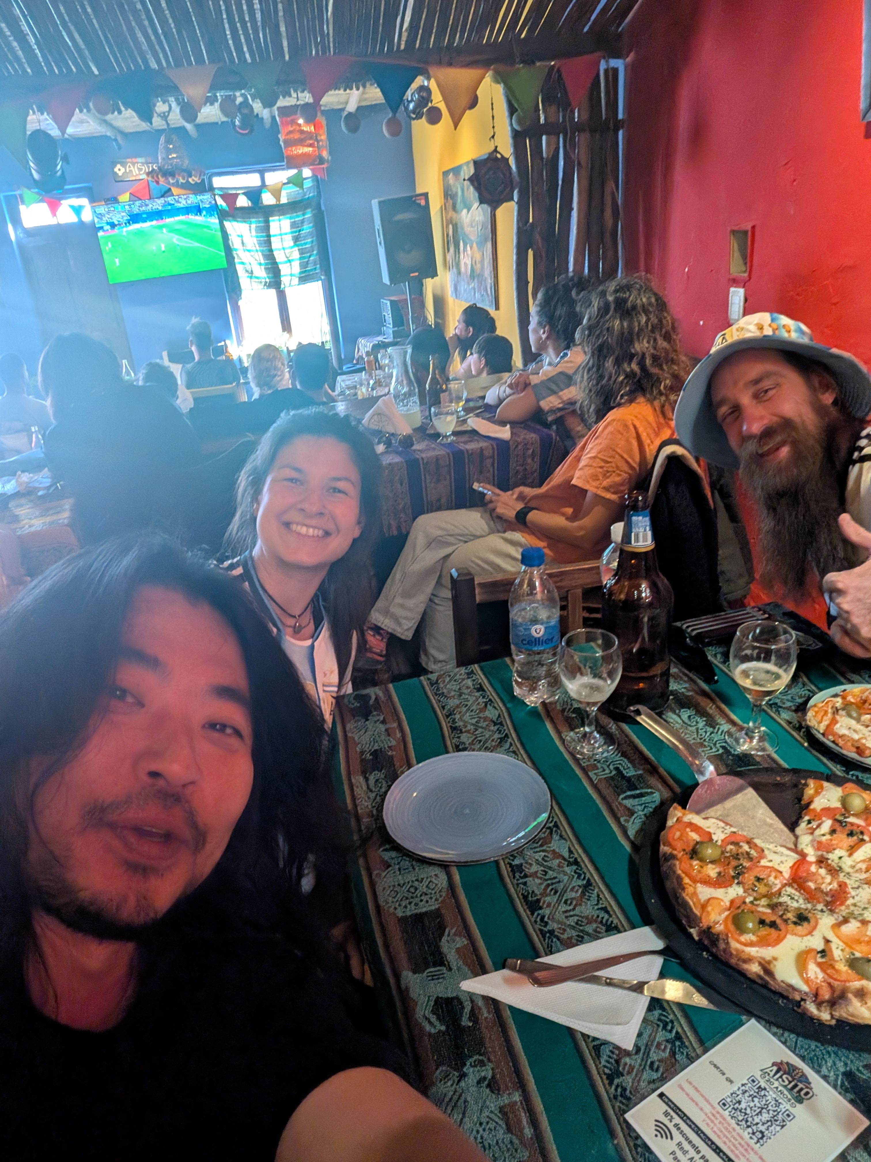 Dorien smiling in a colourful Humahuaca restaurant beside friends she met in Tilcara, with an Argentina football match playing on a screen and pizza and drinks on the table.