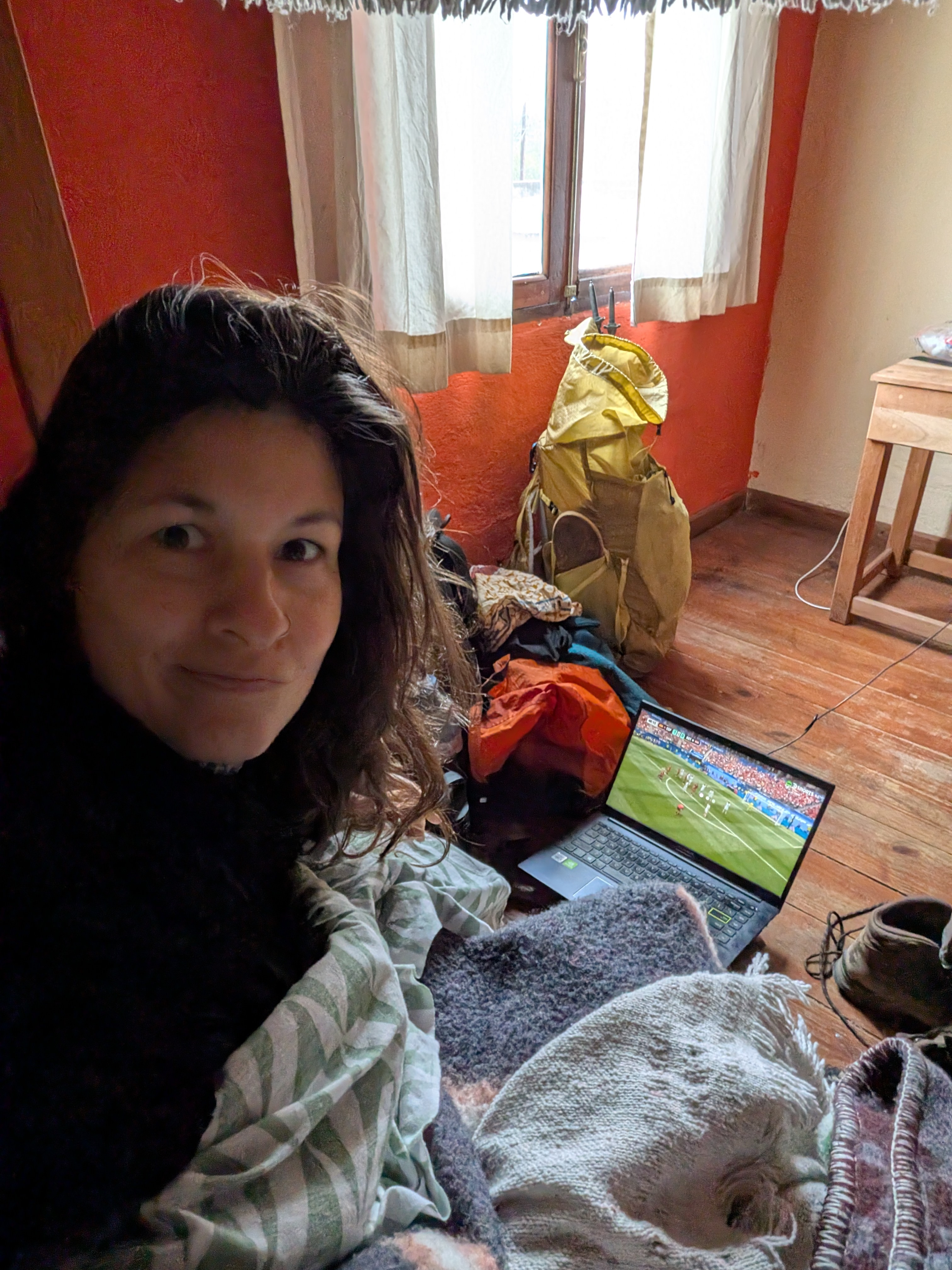 Dorien wrapped in a striped blanket on a bed in a hostel room with warm orange walls, watching a football match on a laptop on the floor, with a backpack and gear beside her.